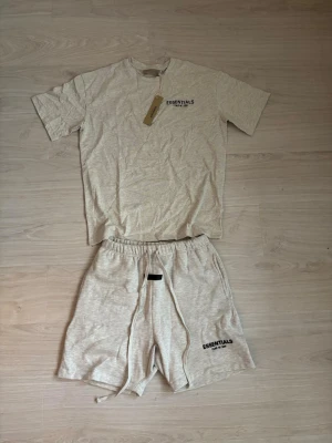 Essentials Fear of God set beige - Säljer ett snyggt set från Essentials Fear of God i ljusbeige. Setet består av en t-shirt med tryck fram och bak samt matchande shorts med dragsko och logga på benet. Materialet är mjukt och bekvämt, perfekt för en avslappnad stil.