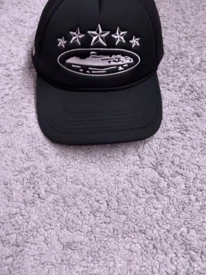 Svart corteiz keps - Snygg svart truckerkeps med vit brodyr framtill som visar fem stjärnor och ett motiv, samt mesh på baksidan för extra ventilation. Justerbar storlek med snapback-spänne och broderad text bak. Perfekt för en streetwear-look.