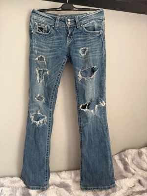 Blå slitna bootcut jeans med nitar - Säljer ett par blå bootcut jeans med  slitningar och hål framtill. Baksidan har detaljerade bakfickor med nitar och mönster. Jeansen har låg midja och klassisk femficksdesign. Perfekta för dig som gillar en edgy och trendig stil.