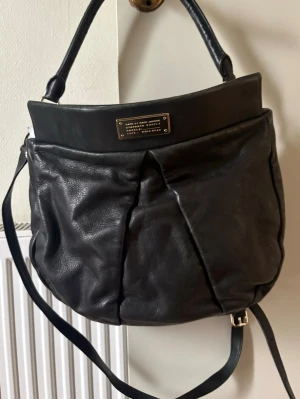 Svart handväska i skinn från Marc Jacobs - Snygg svart handväska i mjukt skinn från Marc Jacobs. Väskan har en rundad form, guldfärgad logoplatta framtill och både axelrem och handtag. Perfekt storlek för att få plats med allt du behöver och passar till många olika stilar. Perfekt till skolan! Läkta läder skriv för fler bilder 🌸