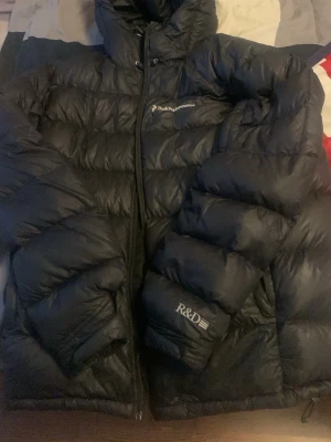 Svart dunjacka från Peak Performance - Säljer en svart dunjacka från Peak Performance med huva och dragkedja. Jackan har broderad logga på bröstet och R&D-detalj på ärmen. Puffig modell som håller dig varm under kalla dagar. Perfekt för dig som vill ha en stilren och funktionell vinterjacka. Dragkedjan har tyvärr gått sönder så denna kan man få till ett bra pris
