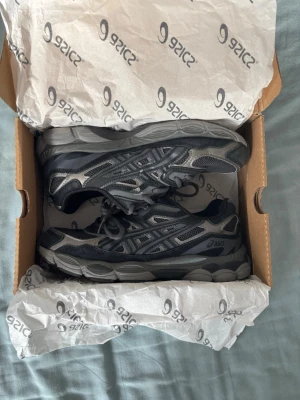 Svarta och grå Asics sneakers - Snygga Asics sneakers i svart och grått med coola detaljer och meshpaneler för extra andningsförmåga. Skorna har en chunky sula och klassisk snörning. Perfekta för dig som gillar sportig och trendig stil.