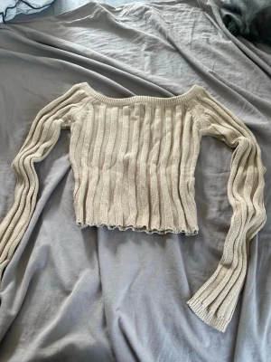 Beige ribbad off shoulder stickad tröja - Trendig beige stickad tröja med ribbad struktur och off shoulder-design. Croppad modell med långa, vida ärmar och vågig kant nertill. Perfekt för dig som gillar en avslappnad men ändå stilren look.