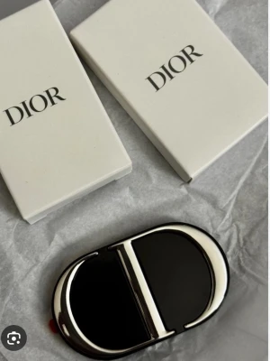 Svart Dior spegel med metallogo - Snyggt svart spegel från Dior med en stor silverfärgad metallogo på baksidan. Skalet har en glansig finish och exklusiv känsla, perfekt för dig som vill ha en lyxig look på mobilen. Passar till iPhone och ger både stil och skydd.