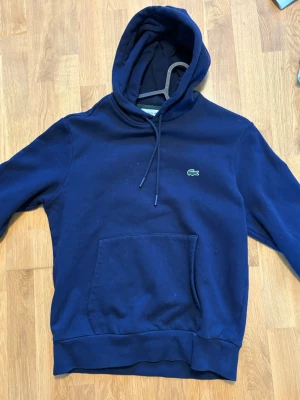 Mörkblå hoodie från Lacoste - Mörkblå hoodie från Lacoste med klassisk krokodillogga på bröstet. Hoodien har huva med dragsko och en stor magficka framtill. Perfekt för en avslappnad och stilren look.