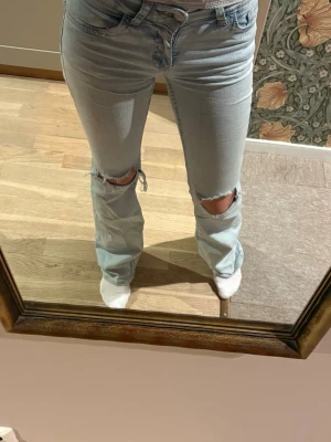 Ljusblå slitna bootcut jeans - Säljer ett par ljusblå bootcut jeans med slitningar över knäna och broderade detaljer på bakfickorna. Jeansen har klassisk femficksdesign och är tillverkade i denim med stretch för skön passform.