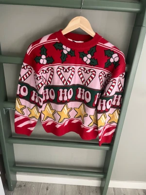 Jultröja med mönster från H&M - Säljer en färgglad stickad jultröja från H&M med röda, vita och gröna detaljer. Tröjan har motiv av mistel, polkagrisar, stjärnor och texten HO HO HO. Perfekt för julmys och att sprida festlig stämning.