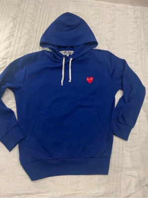 Blå hoodie med hjärtdetalj - Blå hoodie med vit dragsko och en röd hjärtdetalj med ögon broderad på bröstet. Klassisk känguruficka framtill och ribbade muddar vid ärmar och nederkant. Perfekt för en chill och avslappnad stil.