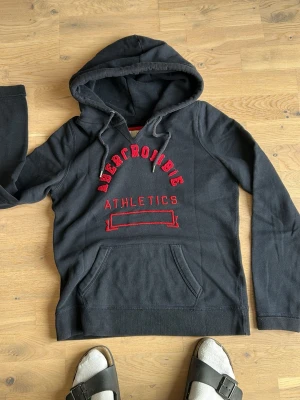 Mörkgrå Abercrombie hoodie - Snygg mörkgrå hoodie från Abercrombie med röd broderad text 'Abercrombie Athletics' på bröstet. Klassisk känguruficka framtill och justerbar huva med snören. Perfekt för en avslappnad och sportig stil.