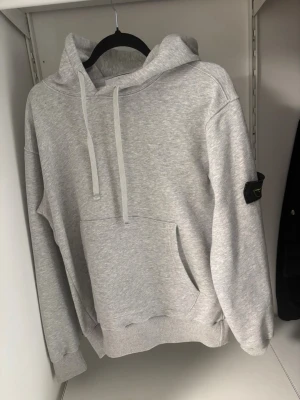 Grå hoodie från Stone Island - Säljer en ljusgrå hoodie från Stone Island med klassisk patch på vänster ärm. Hoodien har huva med dragsko, stor magficka och ribbade muddar vid ärmslut och nederkant. Perfekt för dig som gillar streetwear och vill ha en stilren look.