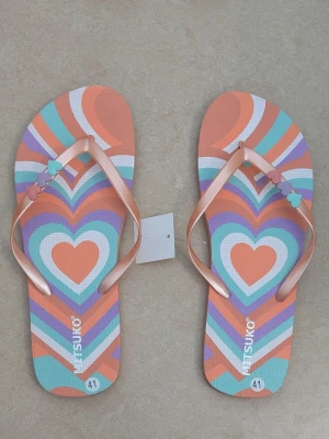 Färgglada hjärtmönstrade flip-flops - Säljer ett par färgglada flip-flops från Mitsuko med hjärtmönster i orange, turkos, lila och vitt. Remmarna är ljusrosa med små hjärtan i olika färger. Perfekta för sommaren och stranden.