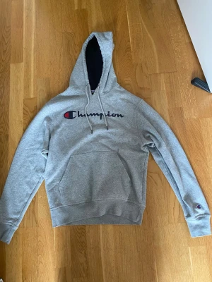Grå Champion hoodie  - Snygg grå hoodie från Champion med stor logga i svart, rött och blått på bröstet. Klassisk känguruficka framtill, dragsko i huvan och ribbade muddar. Perfekt för en avslappnad och sportig stil. Tröjan är i bra skick förutom hålet som syns på sista bilden