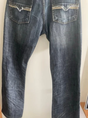 Boots cut jeans diesel  - Säljer ett par blå jeans med slitningar på bakfickorna och tvättad look. Jeansen har raka ben och klassisk femficksdesign. Materialet är denim i bomull och färgen är mörkblå med ljusare partier. Perfekta för en avslappnad och trendig stil.