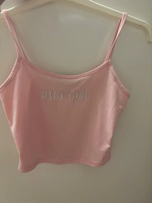 Rosa crop top med texten BABY GIRL - Söt rosa crop top med tunna spaghetti-band och broderad texten 'BABY GIRL' framtill. Tillverkad i mjukt, stretchigt material som sitter bekvämt och ger en trendig look. Perfekt att matcha med höga jeans eller kjol.