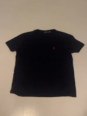 Svart t-shirt från Polo Ralph Lauren - Klassisk svart t-shirt från Polo Ralph Lauren med röd broderad logga på bröstet. T-shirten har rund halsringning och korta ärmar. Perfekt basplagg i mjuk bomull som funkar till allt.