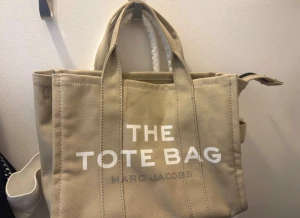 Beige Totebag Marc Jacobs - Snygg beige väska från Marc Jacobs i canvas med trycket 'THE TOTE BAG' på framsidan. Nypris: 2850kr. Väskan har dubbla handtag, praktisk form och detaljerad sömnad. Perfekt för dig som vill ha en trendig och rymlig tote till vardagen.
