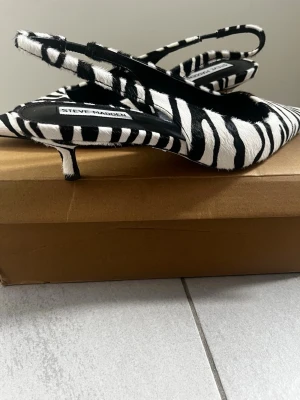 Zebramönstrade pumps från Steve Madden - Säljer ett par trendiga pumps från Steve Madden med zebramönster i svart och vitt. Skorna har spetsig tå, smal klack och öppen häl med rem. Materialet är textil med pälsstruktur för en extra cool look.