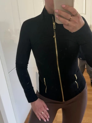Svart jacka från Lululemon med guldzip define nulu jacket  - Stl XS Snygg svart jacka från Lululemon med figurnära passform och guldfärgade dragkedjor framtill och på fickorna. Jackan har hög krage och långa ärmar, perfekt för en sportig och stilren look.