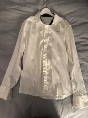 Vit klassisk skjorta med lång ärm - Säljer en stilren vit skjorta med lång ärm och knappar framtill. Skjortan har klassisk krage och en liten broderad detalj på bröstet. Perfekt för en clean och enkel look.