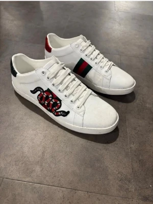 Vita sneakers med orm och ränder - Snygga vita sneakers med broderad röd och svart orm på sidan samt gröna och röda ränder på andra sidan. Skorna har vita snören och detaljer i rött och grönt på hälen. Tillverkade i läder med klassisk låg profil.
