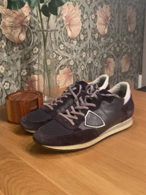 Mörkblå sneakers med mockadetaljer - Snygga mörkblå sneakers med gråa skosnören och vita detaljer. Skorna har en mix av mocka och textilmaterial samt en vit sula med svart kant. På sidan finns en dekorativ patch. Perfekta för en avslappnad och trendig stil.