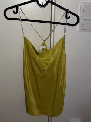  festlig satintopp - Kort gul klänning i satin med tunna, glittriga axelband som korsas i ryggen. Klänningen har en draperad urringning och är ärmlös för en trendig och stilren look. Xs/S