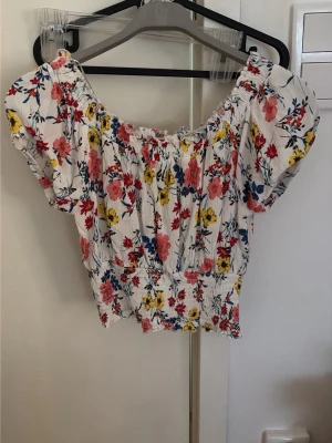 Blommig croppad topp från Selfie - Supersöt croppad topp från Selfie med blommigt mönster i rosa, gul och blå på vit botten. Toppen har korta puffärmar, smockad midja och elastisk halsringning som kan bäras offshoulder. Står M men funkar för S också