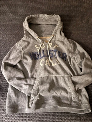 Grå hoodie från Hollister - Säljer en grå hoodie från Hollister med broderad text 'Surf Hollister Cali' i blått och vitt på bröstet. Hoodien har en stor ficka framtill, dragsko i huvan och ribbade muddar. Perfekt för en avslappnad och chill stil. Passar M