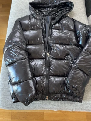 Svart glansig pufferjacka med huva - Säljer en svart, glansig pufferjacka med huva och dragkedja framtill. Jackan har breda kanaler, två sidofickor och elastiska muddar vid ärmsluten. Perfekt för kalla dagar när du vill hålla dig varm och se stilren ut.
