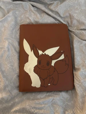 Pokémon pärm med Eevee-motiv - Säljer en brun samlarpärm med Eevee-motiv på framsidan, perfekt för Pokémonkort. Pärmen har plastfickor för att förvara och visa upp korten. Materialet är konstläder och motivet är tryckt i vitt och brunt. Passar dig som vill organisera din samling snyggt.