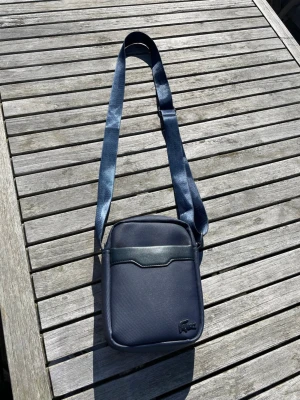 Mörkblå axelväska från Lacoste - Snygg mörkblå axelväska från Lacoste med justerbar axelrem. Väskan har en stilren design med strukturmönstrad yta, diskret Lacoste-logga i hörnet och en dekorativ läderdetalj på framsidan. Perfekt storlek för det viktigaste och enkel att bära över axeln.