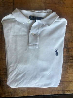 Vit pikétröja från Polo Ralph Lauren - Klassisk vit pikétröja från Polo Ralph Lauren med korta ärmar, krage och två knappar framtill. Tröjan har den ikoniska broderade loggan i mörkblått på bröstet. Perfekt för en stilren och avslappnad look. En av slitsarna på tröjan är lite nött (se bild). Priset är inte hugget i sten