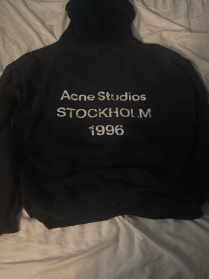 Svart hoodie Acne Studios - Säljer en svart hoodie från Acne Studios med tryck på ryggen där det står 'Acne Studios STOCKHOLM 1996'. Hoodien har klassisk passform, huva och ribbade muddar. Perfekt för dig som gillar stilrena och trendiga plagg.