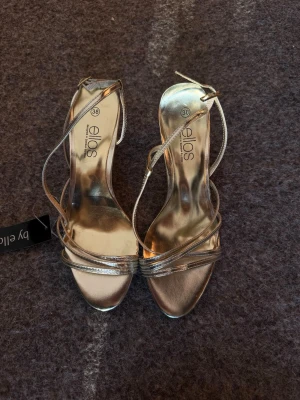 Guldiga pumps med smala remmar - Säljer ett par glansiga guldiga pumps från Ellos med smala remmar och öppen tå. Skorna har en elegant klack och tunna band som korsar foten för en snygg look. Perfekta för att lyfta vilken outfit som helst.
