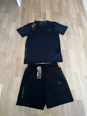 Svart träningsset från Under Armour - Snyggt svart träningsset från Under Armour med t-shirt och shorts. T-shirten har rund hals och korta ärmar, medan shortsen har resårmidja och dragsko. Båda plaggen har diskreta loggor och är tillverkade i ett lätt, snabbtorkande material som passar perfekt för träning.