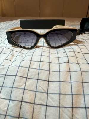 Beige och svarta solglasögon Chanel - Säljer ett par ikoniska solglasögon från Chanel med beige bågar och svarta sidor, prydda med den klassiska Chanel-loggan i guld. Glasen är mörka och modellen har en chunky, retroinspirerad form. Perfekt för dig som vill sticka ut med lyxig accessoar.