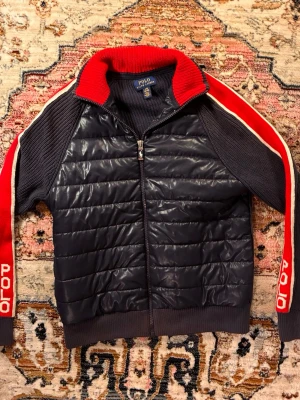 Mörkblå ziptröja från Polo Ralph Lauren - Snygg mörkblå ziptröja med quiltad framsida och stickade ärmar. Röda och vita ränder längs ärmarna med 'POLO' tryckt på båda sidor. Hög krage med röd insida och ribbade muddar. Perfekt för en sportig och avslappnad look.