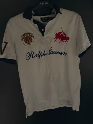 Vit piké från Ralph Lauren - Vit pikétröja från Ralph Lauren med klassisk passform. Stora broderade detaljer på bröstet, inklusive röd polospelare och blå Ralph Lauren-text. Mörkblå krage och ärmslut samt broderad patch och bokstav på ärmen.