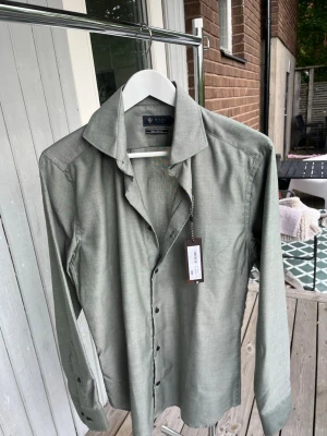 Premium skjorta – Clean & Stilren Look | Perfekt för varje tillfälle - Stilren skjorta i en elegant ljusgrå/grön nyans med klassisk krage och diskreta mörka knappar. Den mjuka och följsamma passformen gör den enkel att bära både till vardags och vid finare tillfällen. Matcha med jeans eller kostymbyxor för en modern, clean look.