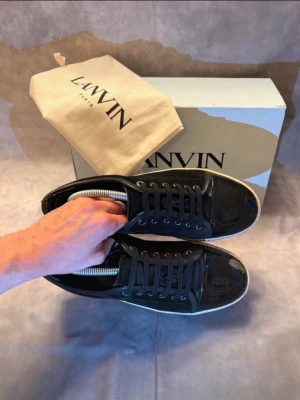 Lanvin skor - Säljer ett par svarta sneakers från Lanvin med klassisk låg profil och vit sula. Skorna har snörning och är tillverkade i en mix av mocka och skinn, vilket ger en snygg kontrast mellan material. Perfekta för dig som gillar stilrena och exklusiva sneakers.