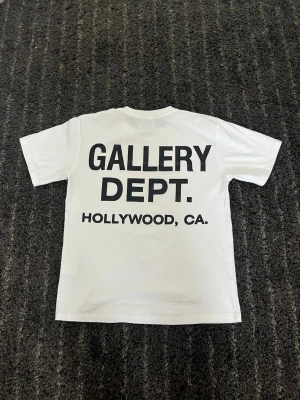 Vit Gallery Dept. t-shirt - Säljer en vit t-shirt från Gallery Dept. med svart tryck fram och stort tryck på ryggen där det står 'GALLERY DEPT. HOLLYWOOD, CA.'. Klassisk passform och rund hals. Perfekt för dig som gillar streetwear och enkel stil.