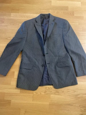 Grå kavaj från Michael Kors - Stilren grå kavaj från Michael Kors med diskret rutmönster. Tvåknäppt modell med klassisk krage, två fickor med lock och en bröstficka. Fodrad insida och snygg passform, perfekt till skjorta och jeans.