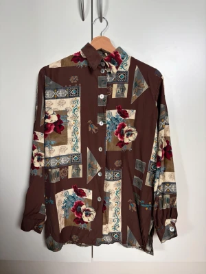 Brun vintage skjorta med blommigt mönster - Brun vintageskjorta med knäppning framtill och slits på sidorna i strl 36. Inga synliga skador. Det står tyvärr inget om material, men den är tunn och upplevs sval & luftig. Vääääääääldigt minimal see-through men när man har på sig den så ser man verkligen inte igenom (se bild 7 där jag håller upp den mot ljuset från fönstret)