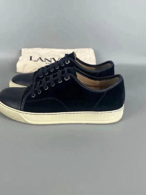 Blåa Lanvin sneakers i mocka - Snygga blåa sneakers från Lanvin med ovandel i mocka och detaljer i läder. Skorna har vita sulor och klassisk låg profil med svarta skosnören och metallöljetter. Perfekt för dig som gillar stilrena och exklusiva sneakers.