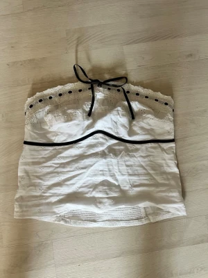 Vit axelbandslös spets tubtopp - Söt vit axelbandslös tubtopp med broderade detaljer och spetskant upptill. Svart band med rosett framtill ger en snygg kontrast. Perfekt till sommaren och enkel att matcha med jeans eller kjol.