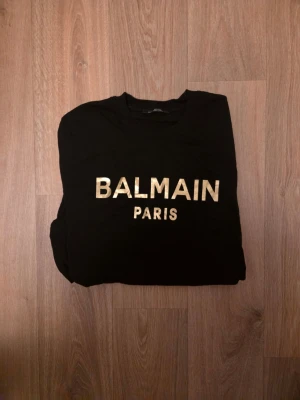 Svart Balmain Paris t-shirt - Snygg svart t-shirt från Balmain med guldig text 'BALMAIN PARIS' på bröstet. Klassisk passform med rund hals och korta ärmar. Perfekt för dig som vill ha en stilren och trendig look.
