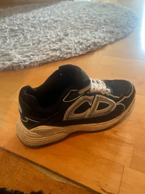 B30 - Snygga svarta sneakers med vita detaljer och tjock vit sula. Skon har ett stort CD-tryck på sidan och vita skosnören. Ovandelen är i textil och syntetmaterial, perfekt för en trendig streetstil.