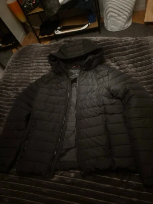 Superdry jacka   - Säljer en svart pufferjacka med huva och dragkedja framtill. Jackan har quiltad design med horisontella sömmar och två fickor med dragkedja. Perfekt för alla årstider och riktigt skön att bära. Materialet är syntet och jackan är vadderad för extra värme. 