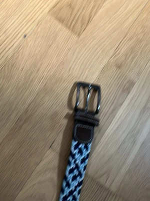 Blåvitt flätat bälte med läderdetalj - Snyggt flätat bälte i blått och vitt med läderdetalj vid spännet. Bältet har ett randigt mönster och metallspänne, perfekt för att ge din outfit en cool touch.