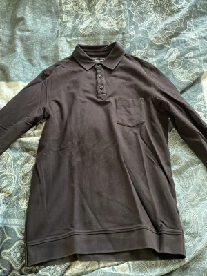 Mörkbrun pikétröja från Dressmann - Snygg mörkbrun långärmad pikétröja från Dressmann med klassisk krage, tre knappar och bröstficka. Tillverkad i mjukt bomullstyg som känns skönt mot huden. Perfekt för en avslappnad men stilren look.